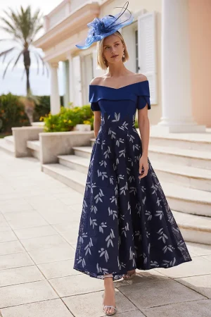 A woman wears the Rosa Clara - 9G1F3 off-shoulder blue dress and hat, standing outdoors near a pastel building with palm trees.