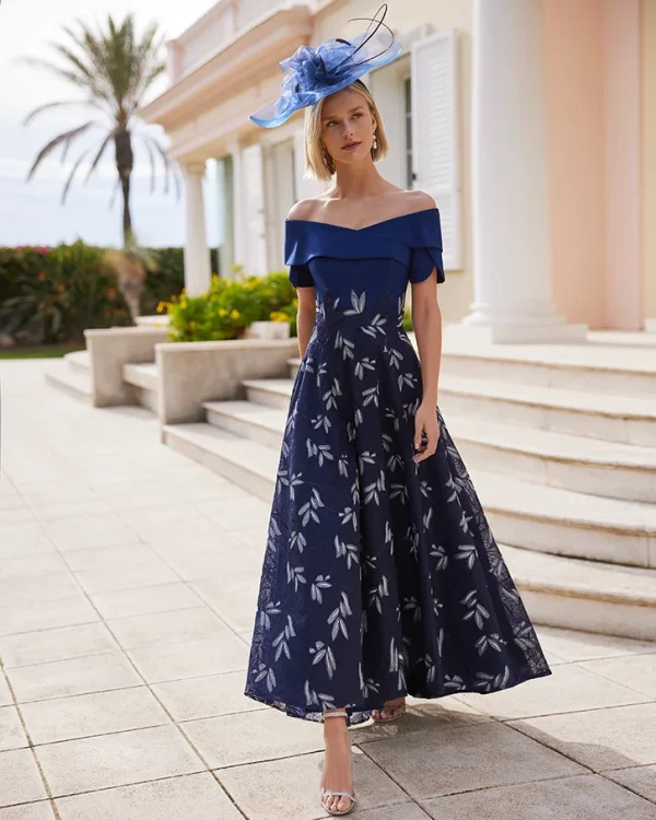 A woman wears the Rosa Clara - 9G1F3 off-shoulder blue dress and hat, standing outdoors near a pastel building with palm trees.