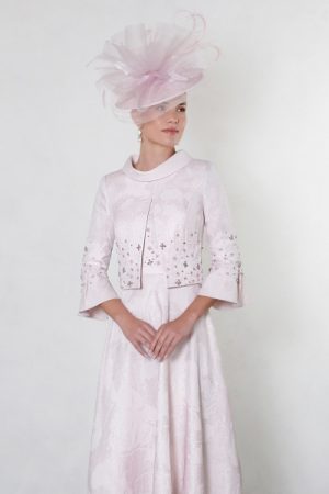 Woman wearing the Rosa dress in pale pink with a matching hat featuring tulle detail, standing against a plain white background.