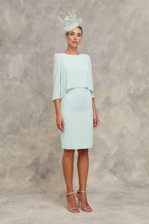 Claudia C - Begur showcases elegance with a light green knee-length dress and matching fascinator, set against a neutral backdrop—ideal for sophisticated Begur events.
