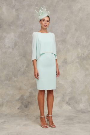 Claudia C - Begur showcases elegance with a light green knee-length dress and matching fascinator, set against a neutral backdrop—ideal for sophisticated Begur events.