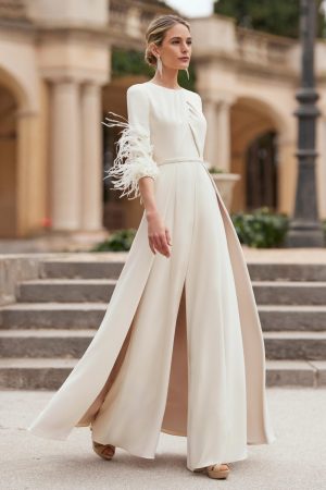Woman wearing the Rosa Clara - 8G145 ivory jumpsuit with feathered sleeves, walking outdoors near stone steps and columns.