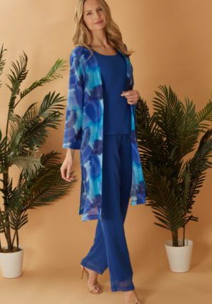 A woman wearing Glitz - 9985, a blue outfit with a floral sheer robe, stands between two potted plants against a tan background.