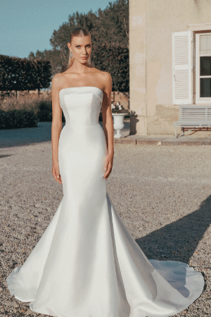 A woman in the Justin Alexander 44456 - Paris strapless white gown poses gracefully outdoors near a shuttered building on a sunny day, capturing Parisian elegance.