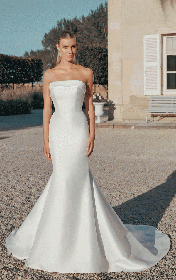 A woman in the Justin Alexander 44456 - Paris strapless white gown poses gracefully outdoors near a shuttered building on a sunny day, capturing Parisian elegance.