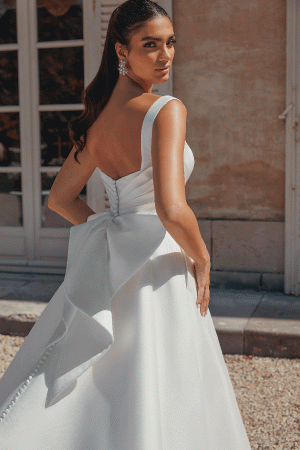 In a Justin Alexander 44457 - Bronte gown, featuring a large bow, a bride poses outside with timeless elegance, glancing over her shoulder.