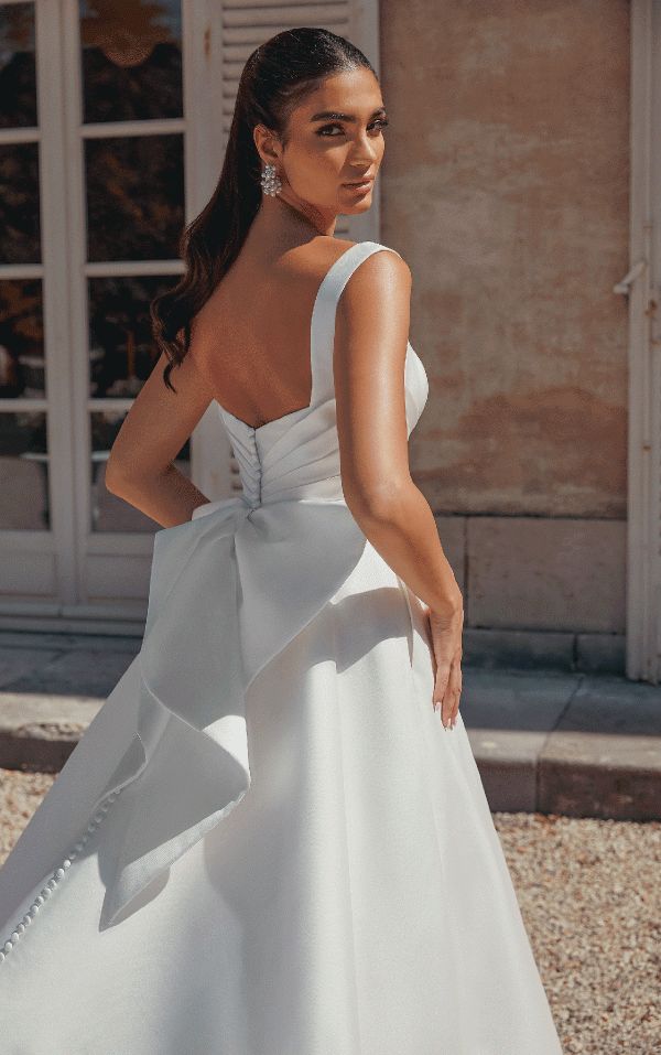 In a Justin Alexander 44457 - Bronte gown, featuring a large bow, a bride poses outside with timeless elegance, glancing over her shoulder.