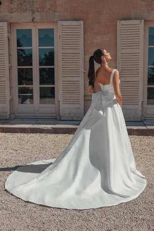 In a Justin Alexander 44457 - Bronte gown with a flowing train, the bride stands outside by white shutters, gazing up wistfully.