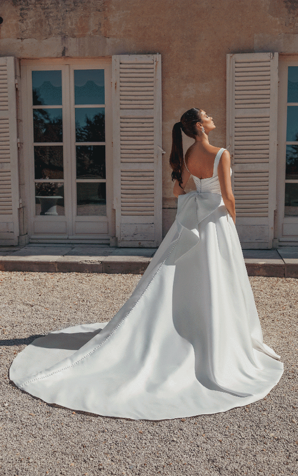 In a Justin Alexander 44457 - Bronte gown with a flowing train, the bride stands outside by white shutters, gazing up wistfully.