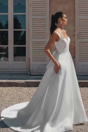 A woman in a white Justin Alexander 44457 - Bronte wedding dress stands outside by a building with wooden shutters, capturing a moment straight out of Bronte's novels.