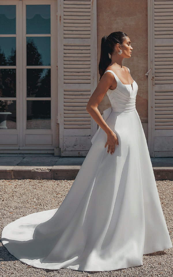 A woman in a white Justin Alexander 44457 - Bronte wedding dress stands outside by a building with wooden shutters, capturing a moment straight out of Bronte's novels.
