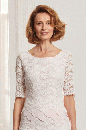Woman with short blonde hair wears the Condici 71125 white lace dress, standing and smiling in a bright room—ideal for special occasions.