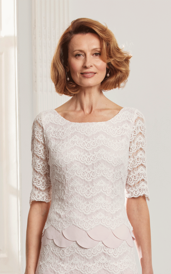 Woman with short blonde hair wears the Condici 71125 white lace dress, standing and smiling in a bright room—ideal for special occasions.