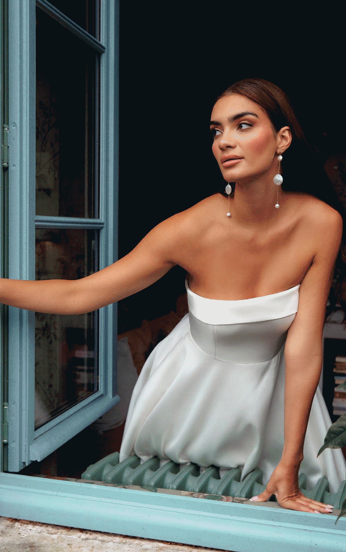 Manon leans gracefully out of a blue window in the strapless white Justin Alexander 44354 - Manon dress, adorned with long pearl earrings.