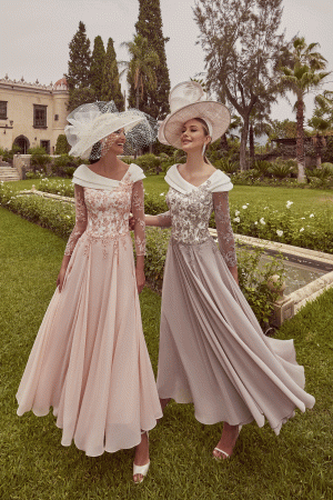 Two women in Veni Infantino - 992423 long dresses and large hats smile at each other while standing in a garden.