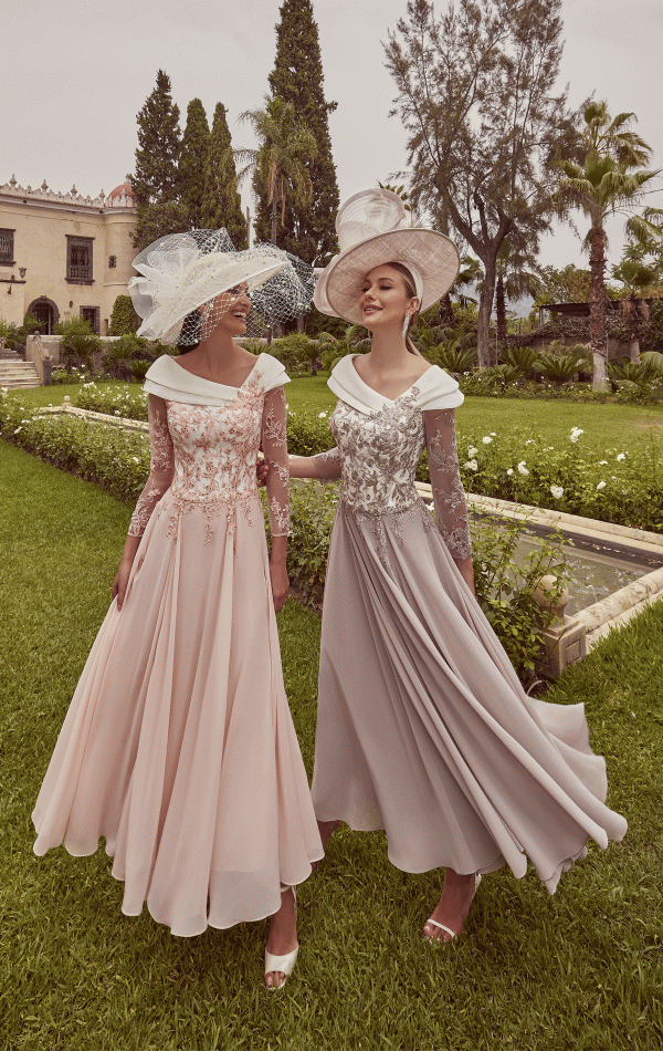 Two women in Veni Infantino - 992423 long dresses and large hats smile at each other while standing in a garden.