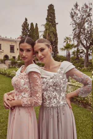 Two women wearing Veni Infantino - 992423 gowns pose together in a lush garden with tall trees and a stately building in the background.