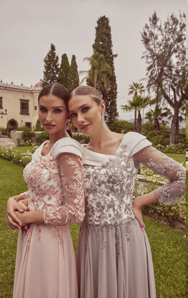Two women wearing Veni Infantino - 992423 gowns pose together in a lush garden with tall trees and a stately building in the background.