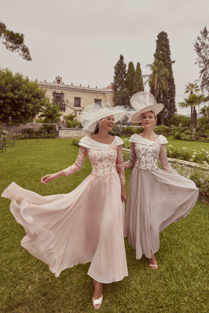 Two women, smiling and walking on grass with a historic building behind them, wear elegant flowing Veni Infantino - 992423 dresses and stylish hats.