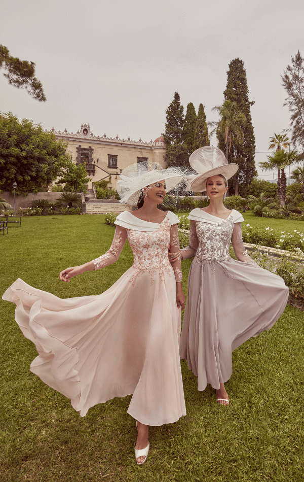 Two women, smiling and walking on grass with a historic building behind them, wear elegant flowing Veni Infantino - 992423 dresses and stylish hats.