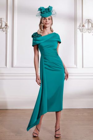 Woman indoors wearing the John Charles - 29291 teal asymmetrical dress with floral shoulder detail, paired with a matching floral hat.