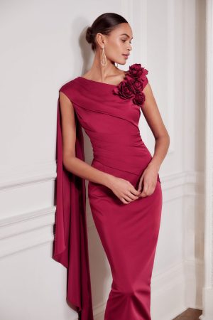 Woman in a fitted magenta John Charles 29278 gown featuring floral shoulder detail and a draped sleeve, standing by a white wall.