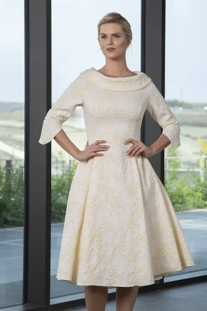 A woman in a cream floral-patterned, knee-length Lizabella 2002 dress stands by large scenic windows—available exclusively for online purchase.