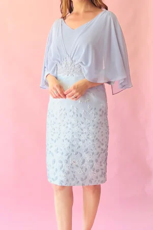 Woman in a light blue Lizabella 2587 dress with floral details, matching hat, and nude heels stands against a pink background—Lizabella 2587 is available for online purchase only.