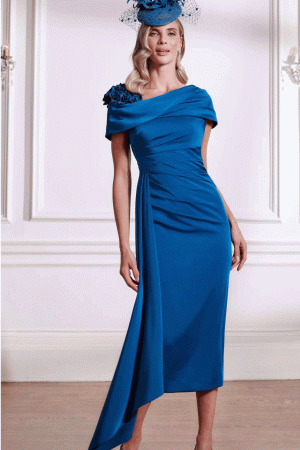 Woman in John Charles - 29291 Teal mid-length dress with ruched bodice and 3D floral shoulder detail, paired with a matching blue floral headpiece. Available at Blessings special occasionwear Brighton.