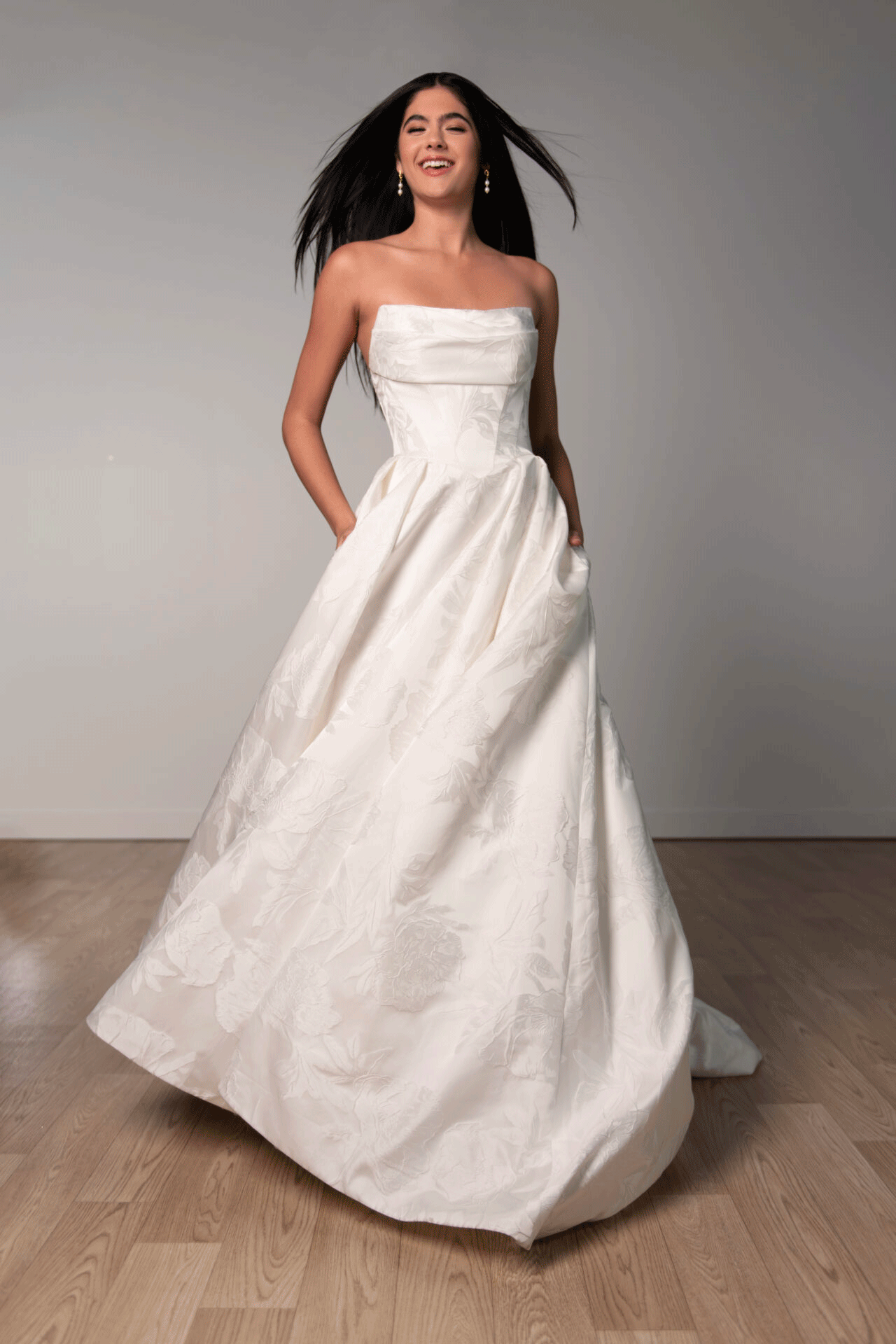 Woman in a strapless white Stella York SY8084 - Ruby wedding gown, smiling and standing on a wooden floor against a plain background.