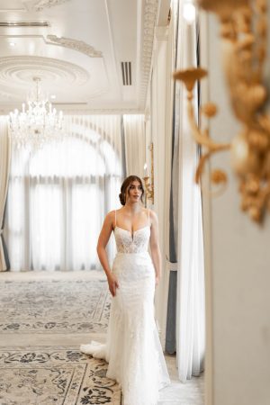 A bride in a Stella York 7863 - Lois white lace wedding dress stands in an elegant, ornate room with chandeliers and large windows.