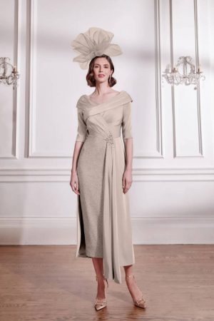 Woman in a beige John Charles - 29289 formal dress with sash detail and a matching large hat, standing in an elegant room.