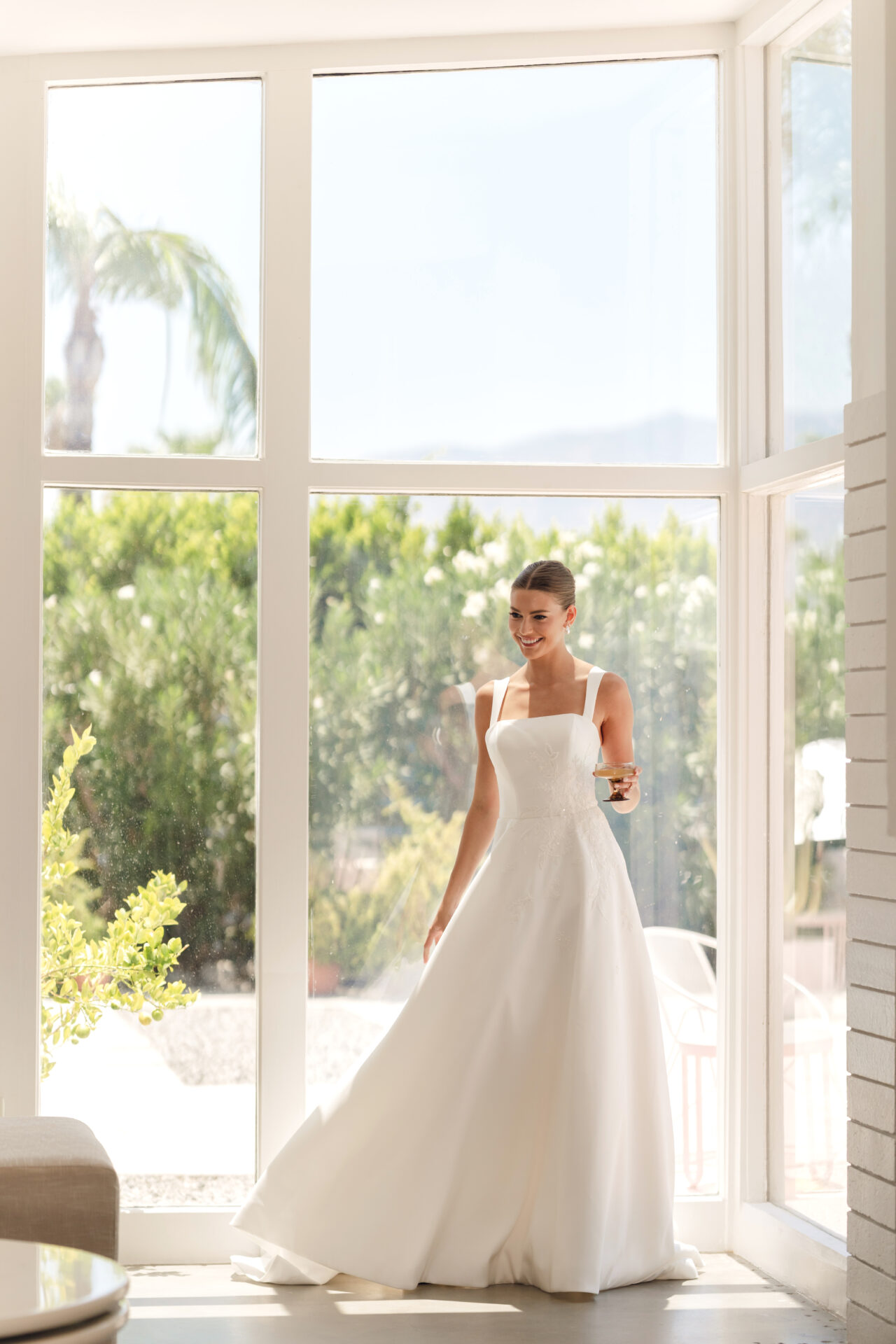 Stella York sy8217 A bride in a white Stella York SY8217 - Daisy wedding dress stands by sunny windows, holding a glass and smiling.