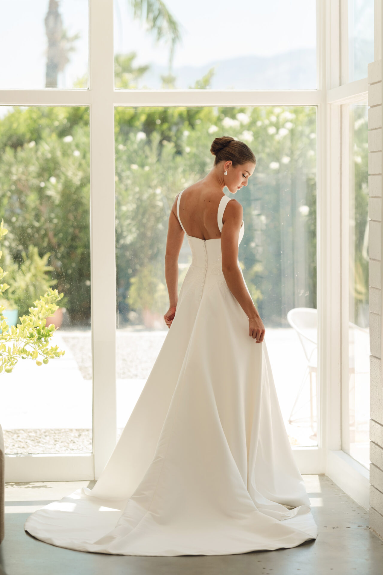 Stella A woman in the Stella York SY8217 - Daisy wedding dress stands by large windows, looking down with her back to the camera.