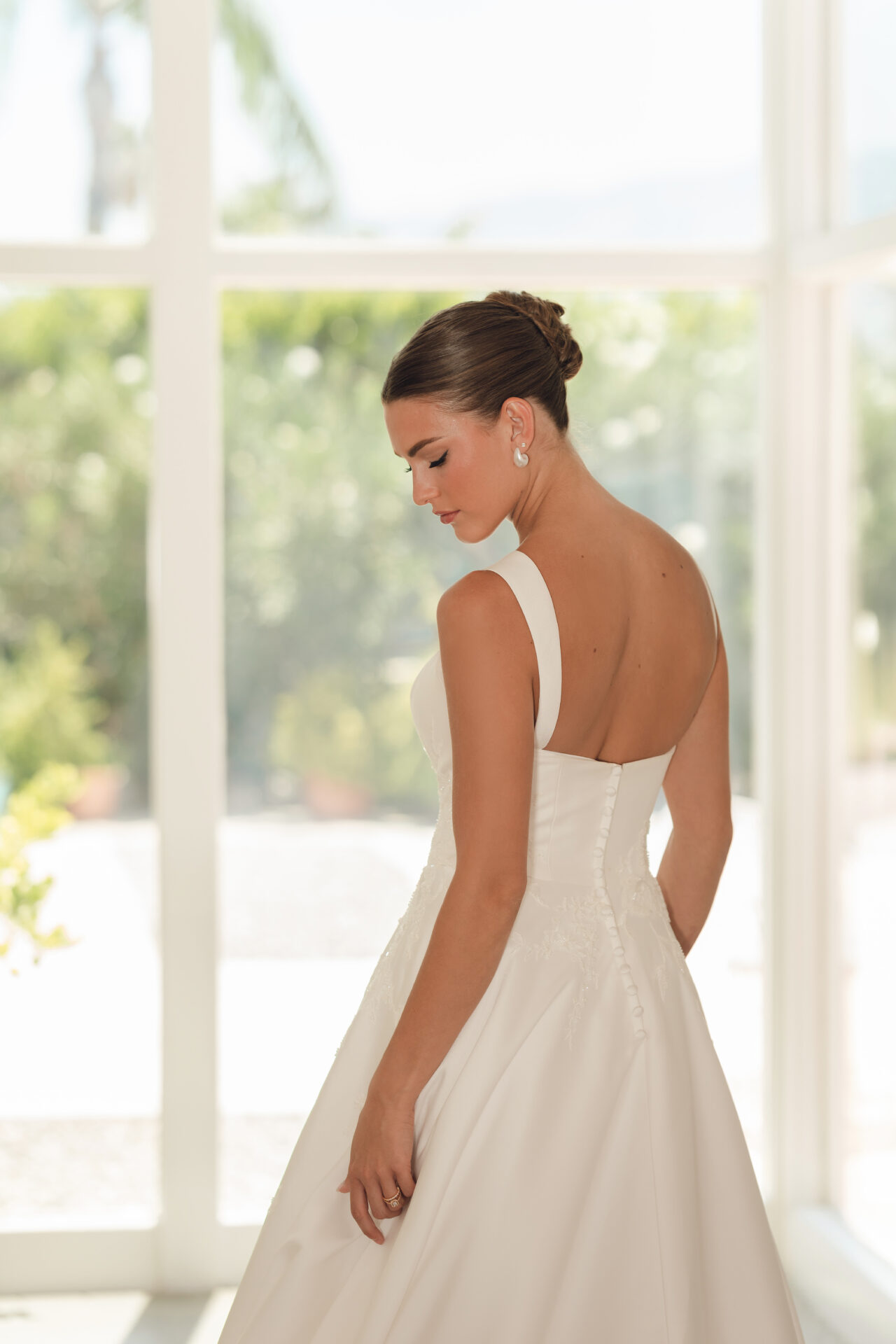 Stella A woman in the Stella York SY8217 - Daisy wedding dress stands indoors, gazing down as sunlight pours through large windows.