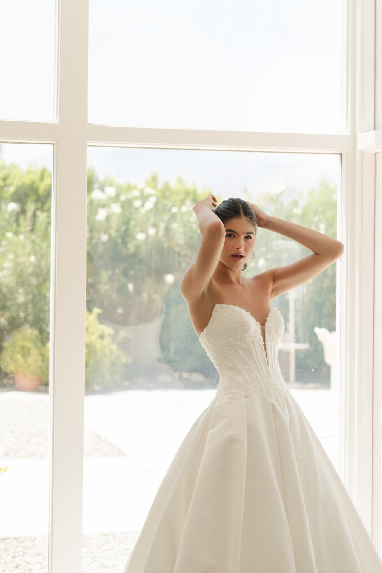 A woman in a strapless white Stella York SY8246 - Annais wedding dress stands by a large window, adjusting her hair.