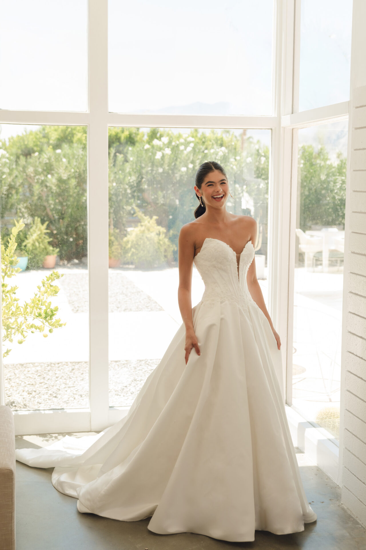 A smiling bride in a strapless ivoryStella York SY8246 - Annais wedding dress stands by large windows with greenery outside. This Satin dress with a basque waist is from Blessings bridal boutique in Brighton