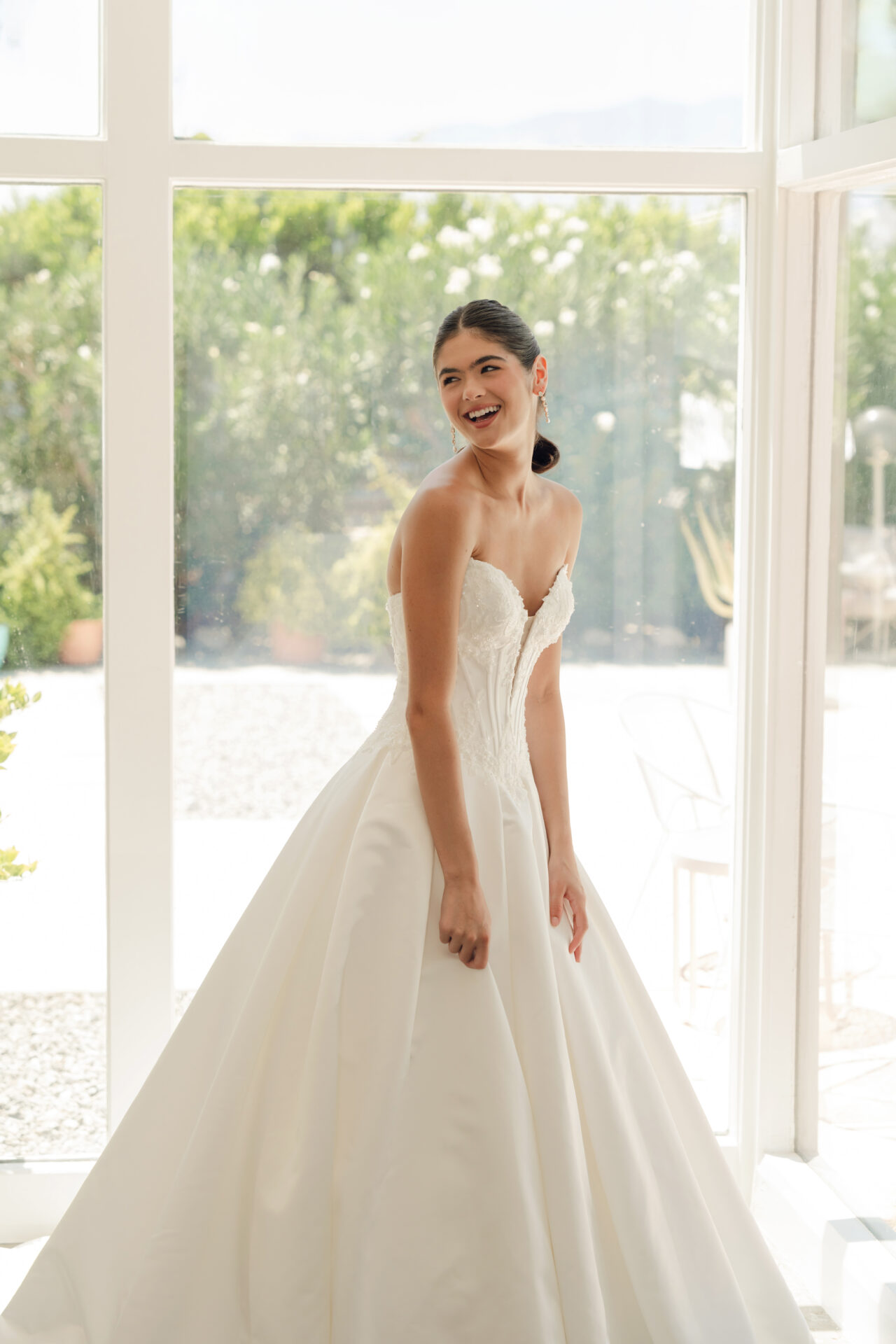A smiling woman in a strapless white Stella York SY8246 - Annais wedding dress stands by a large window with greenery outside.