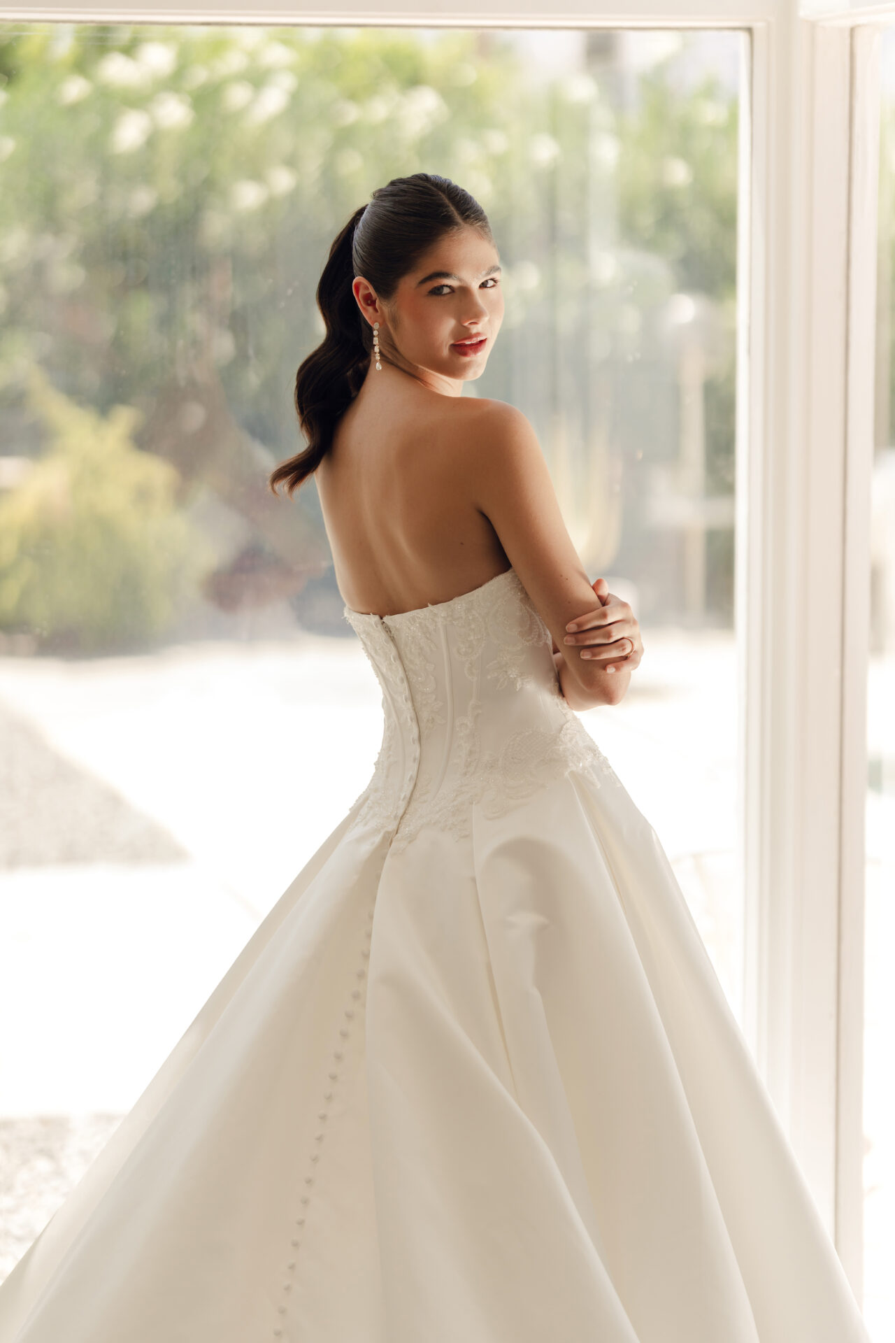 A woman in a strapless white Stella York SY8246 - Annais wedding gown stands indoors, looking over her shoulder at the camera.