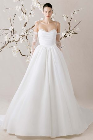 Woman in a white Morilee 1010003 - Èlevè ball gown wedding dress with lace gloves, standing before blossoming branches.
