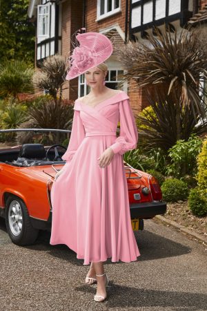 Veni Infantnio 29677: A woman in a pink dress and hat stands beside an orange convertible in front of a house surrounded by plants.