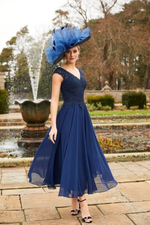 A woman in a flowing blue Veni Infantino - 991905 dress and large matching hat stands by a fountain in a formal garden, radiating elegance.