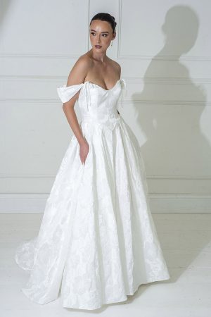 A woman in a Freda Bennet - Meryl off-the-shoulder white wedding gown stands against a white paneled wall, looking to the side.