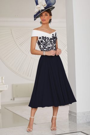 A woman in the Veni Infantino 29516 navy and white off-shoulder designer dress with a large hat stands indoors on a marble floor.