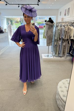 Woman wearing a Veni Infantino - 29651b purple dress and matching hat takes a mirror selfie in a boutique surrounded by formal dresses.