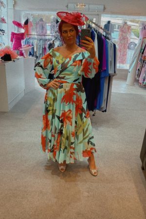 Woman wearing the Veni Infantino - 29826b floral dress and a pink hat takes a mirror selfie in a boutique, surrounded by elegant dresses.