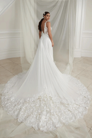 Elyse wears the Justin Alexander 88279 wedding gown, size 12, in white lace with a long ornate train, gracefully posing before sheer curtains. Price: £795.