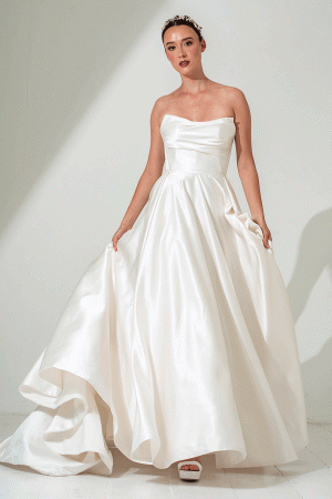 Woman in a strapless, satin, white ballgown-style wedding dress standing against a plain white background.