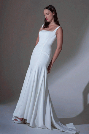Woman wears the Freda Bennet Beau fitted white sleeveless gown with flared train, size 12 (£849), against a plain background. Discover this dress at Blessing of Brighton wedding dress shop in East Sussex.