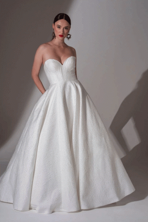 A woman in the Freda Bennet Tyler ball gown, Size 18 (£849), with a strapless sweetheart neckline and textured white fabric, stands against a plain backdrop.